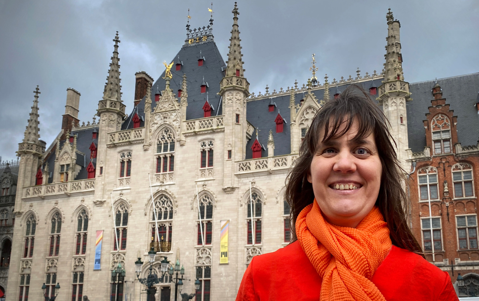 Monique poseert glimlachend voor het stadhuis van Brugge in een oranje outfit